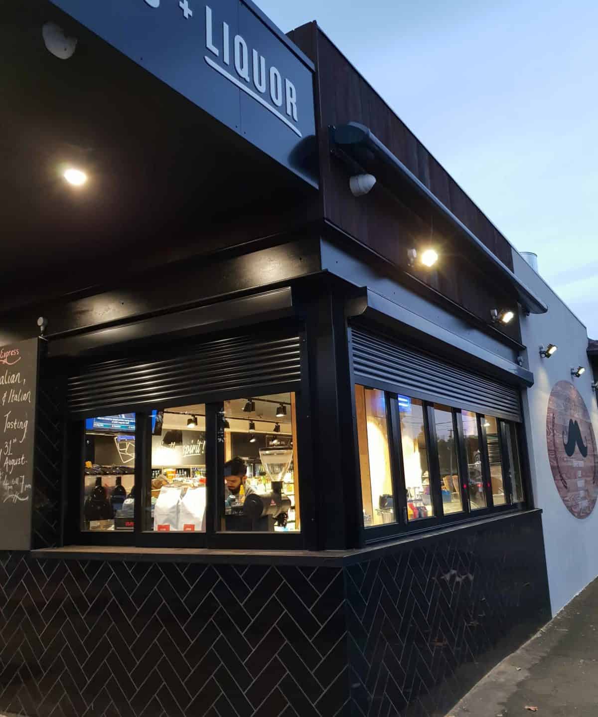 Two charcoal roller shutters partially raised over the shopfront windows of a liquor store, with the interior lit and staff visible behind the glass at dusk.
