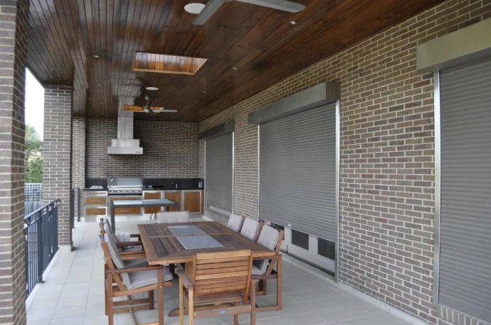 Three silver roller shutters partially lowered along the open side of a large brick alfresco entertaining area, with a timber dining setting, outdoor kitchen with rangehood, and timber-lined ceiling with ceiling fan above.