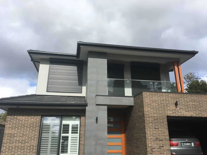 A grey roller shutter partially open on the upper storey of a contemporary two-storey brick home with glass balustrade balcony.