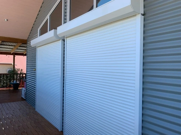 Two white roller shutters fully closed over windows on a home exterior, installed above existing blinds on a timber deck