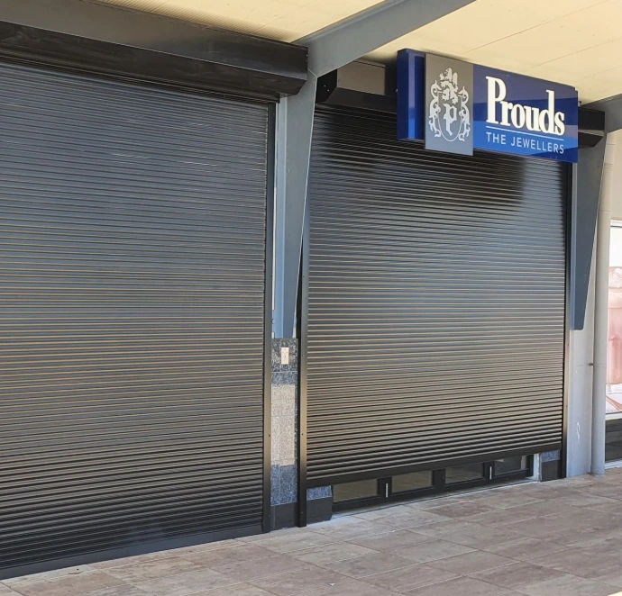 A close-up of a charcoal solid defender security roller shutter closed over the entrance of Prouds The Jewellers, with the branded blue and grey signage prominently displayed above.