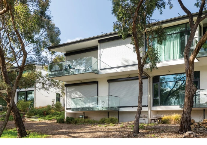 White FireShield roller shutters partially lowered on the upper and lower balconies of a contemporary two-storey rendered home, surrounded by native eucalyptus trees.