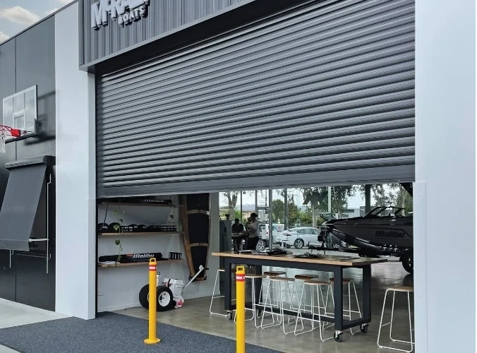A large charcoal SupaSpan roller shutter partially raised over the wide entrance of a Malibu Boats showroom, revealing a bar-height table, stools, and a boat inside.