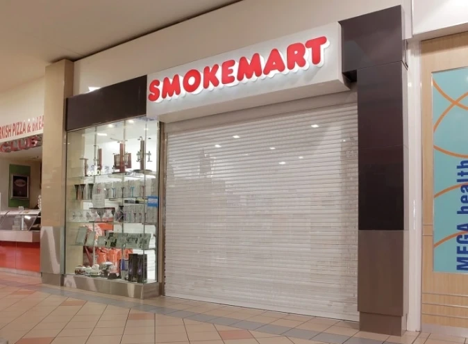 A white extruded vision roller shutter partially closed over half the shopfront of a Smokemart retail store in a shopping centre, with display shelving visible through the open side.