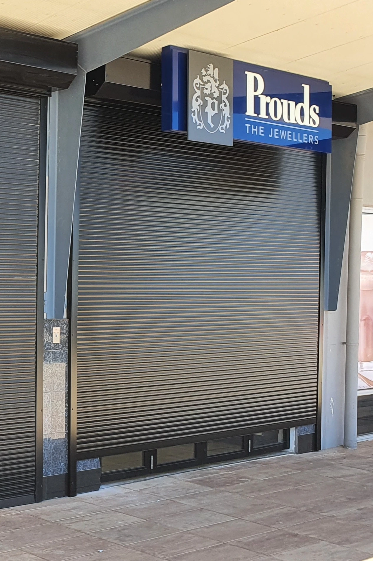 A charcoal solid defender security roller shutter fully closed over the shopfront of Prouds The Jewellers retail store, with the branded blue signage visible above.