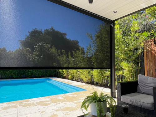 Multiple cream zip-track outdoor blinds fully lowered around a pergola enclosing a wicker outdoor dining setting on a timber deck.