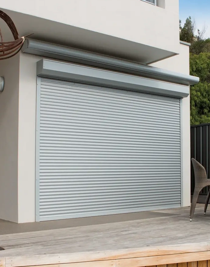 A large silver widespan roller shutter fully closed over a wide opening on the deck of a modern two-storey rendered home, with a rattan chair and timber decking visible.
