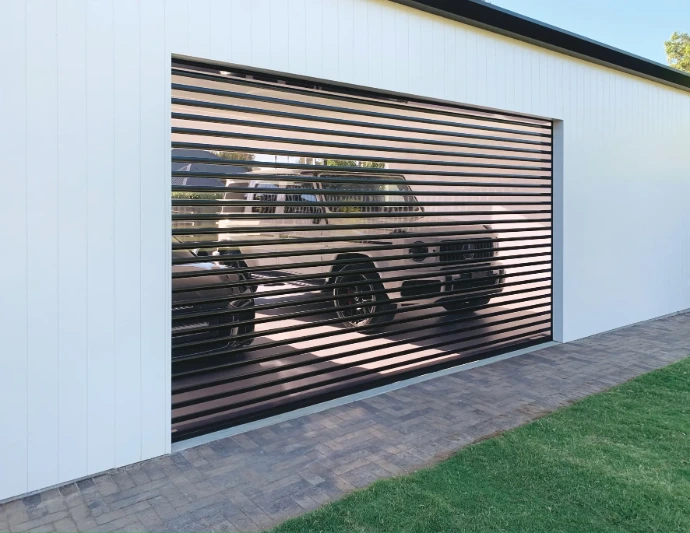 A charcoal clearview louvred roller shutter installed as a garage door on a white clad shed, with a four-wheel drive vehicle silhouetted through the open slats and a paved driveway below.