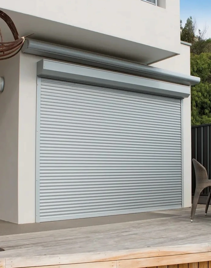 A large roller shutter fully closed over a wide opening on the deck of a modern two-storey rendered home, with a rattan chair and timber decking visible