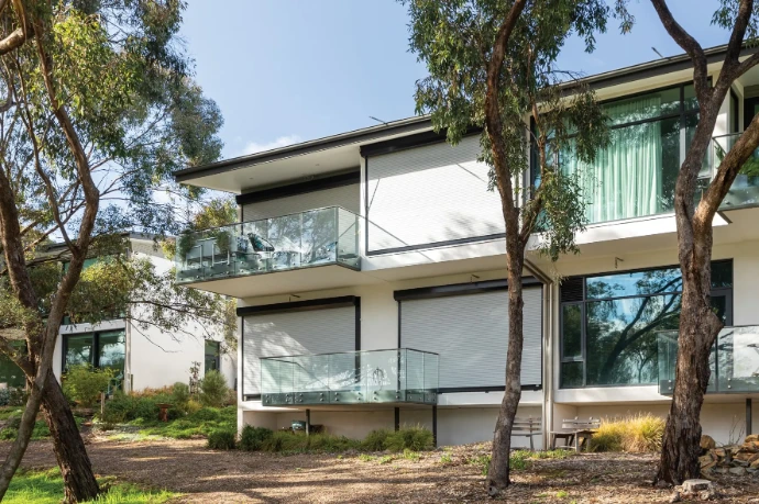 White FireShield roller shutters partially lowered on the upper and lower balconies of a contemporary two-storey rendered home, surrounded by native eucalyptus trees.