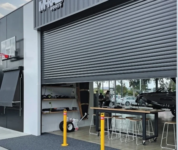 A large charcoal SupaSpan roller shutter partially raised over the wide entrance of a Malibu Boats showroom, revealing a bar-height table, stools, and a boat inside.