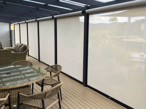Multiple cream zip-track outdoor blinds fully lowered around a pergola enclosing a wicker outdoor dining setting on a timber deck.