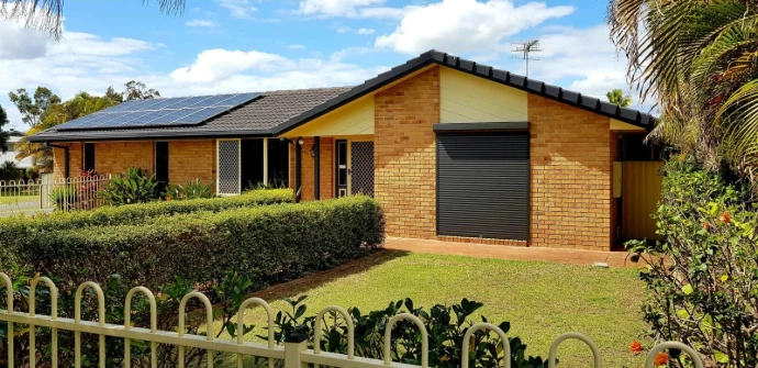 A single charcoal roller shutter fully closed over a window on the front of a tan brick home, with solar panels on the roof, hedge screening, and lush garden surrounds.