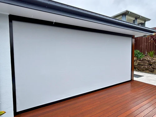 A cream roller shutter fully closed over a large window on a red brick home exterior, set in a charcoal aluminium frame with garden foliage in the foreground.
