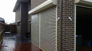 A white louvred clearview roller shutter partially open on the side wall of a double-storey dark brick home, with a security light visible above.