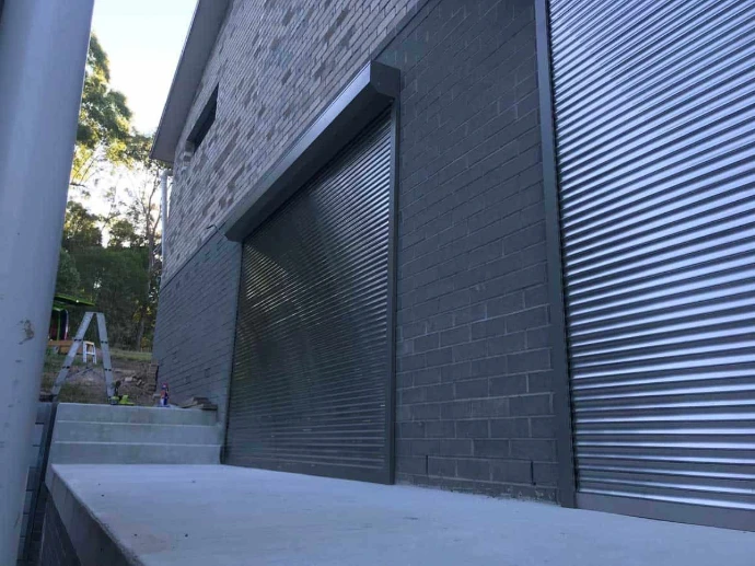 Two large charcoal roller shutters installed on the exterior of a modern dark brick building, photographed from ground level during construction.