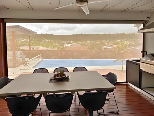 A translucent zip-track outdoor blind partially lowered on a covered alfresco dining area with a pool and suburban rooftop views beyond, ceiling fan above.