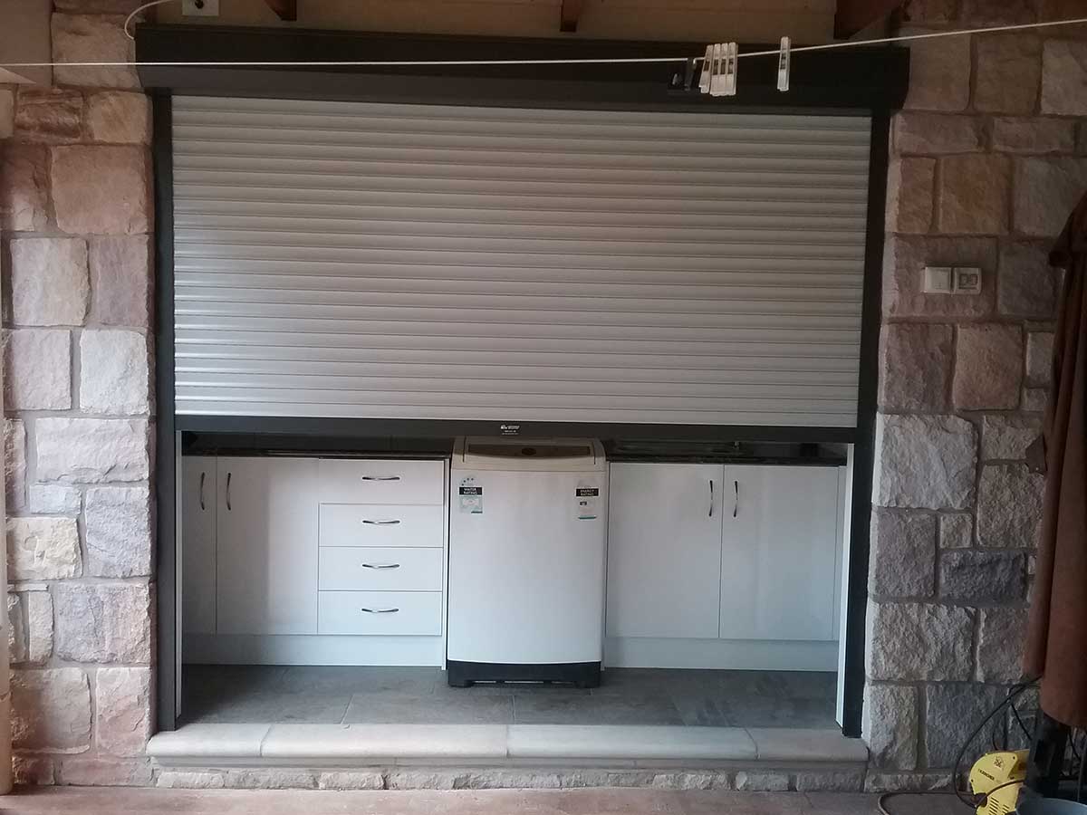 A white roller shutter partially raised over an outdoor laundry alcove set within a limestone block wall, revealing cabinets, drawers, and a top-load washing machine beneath.