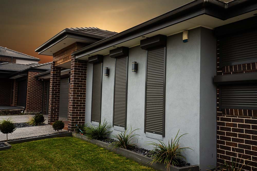 Three charcoal roller shutters fully closed over windows on the facade of a modern rendered and brick home, photographed at dusk with a dramatic warm sky.