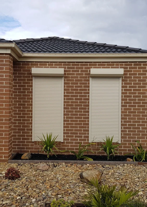 Two cream roller shutters fully closed over side-by-side windows on a red brick home exterior, with garden plants and a gravel garden bed in the foreground.