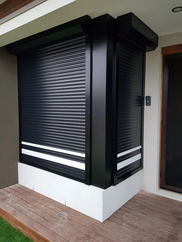 A large black roller shutter partially closed over a corner window bay on a rendered home, with a timber deck and security intercom panel beside the entry door.