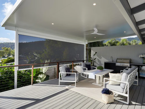 A charcoal zip-track outdoor blind partially lowered on a raised deck alfresco with white outdoor lounge furniture, BBQ, and treetop views beyond.