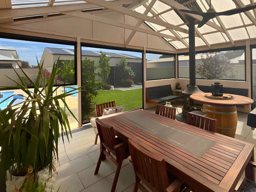Dark insect-screen outdoor blinds enclosing a gabled conservatory-style alfresco with timber dining table, wood barrel, and pool views, photographed from inside.