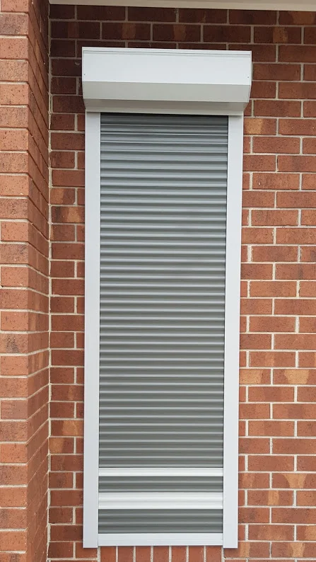 A single silver roller shutter fully closed over a tall narrow window on a red brick home exterior, with a white pelmet box above.