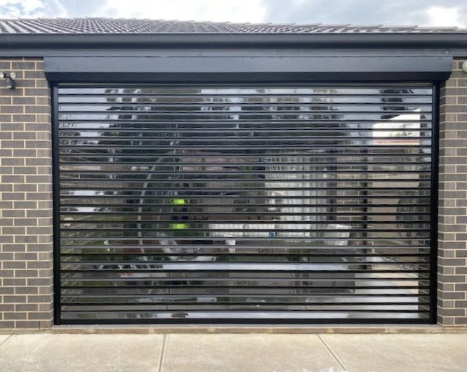A dark charcoal clearview louvred roller shutter installed as a garage door on a tan brick home, with reflections of surrounding trees visible through the slats.