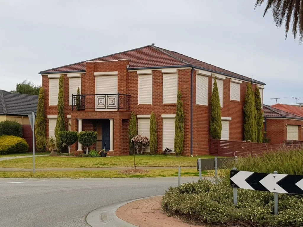 A two-storey red brick home with multiple cream roller shutters installed across all ground and first floor windows, photographed from the street at a roundabout.