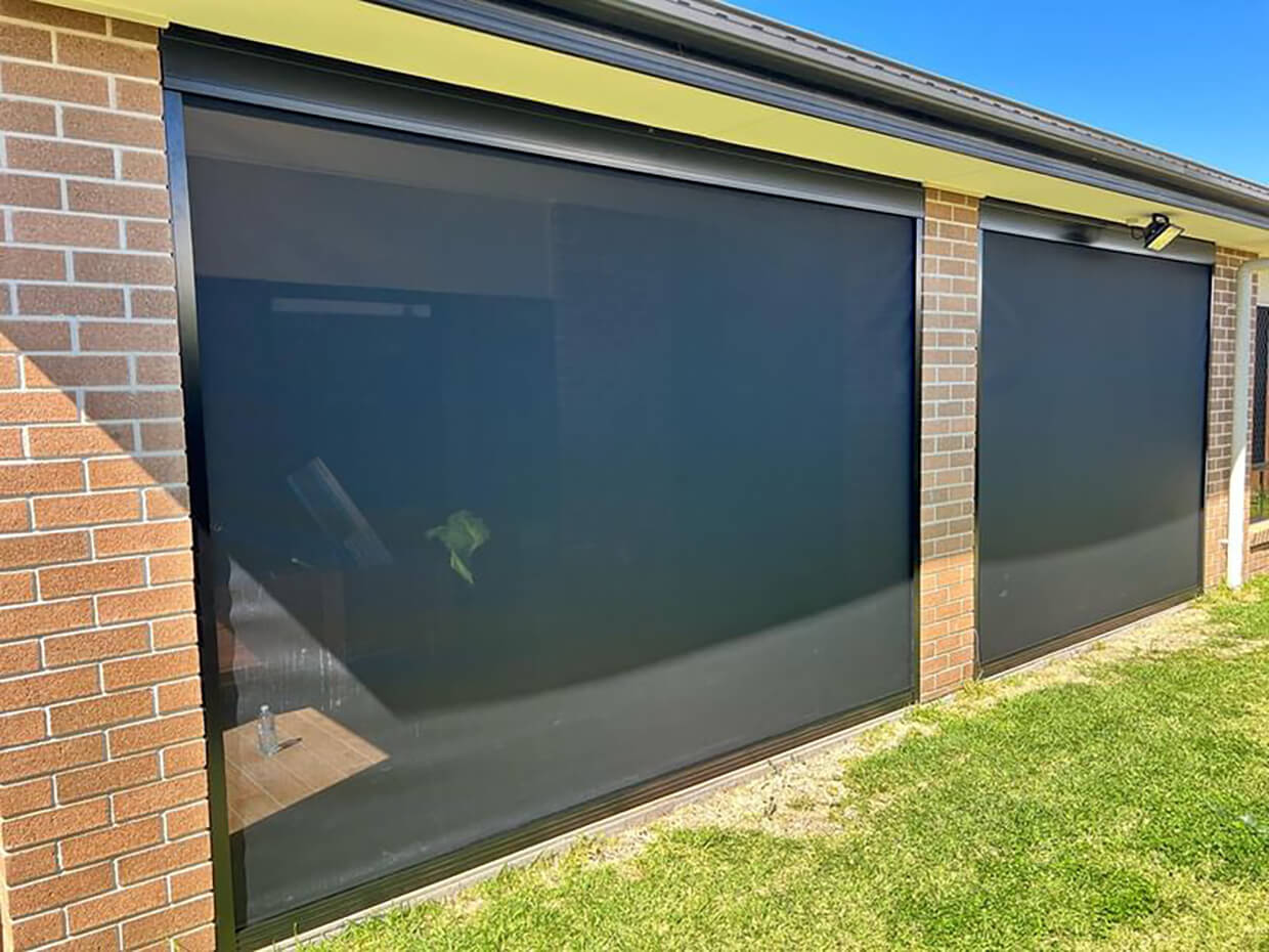 Two large charcoal slide-track outdoor blinds fully lowered across the side wall of a red brick alfresco, with lawn and a security floodlight visible.