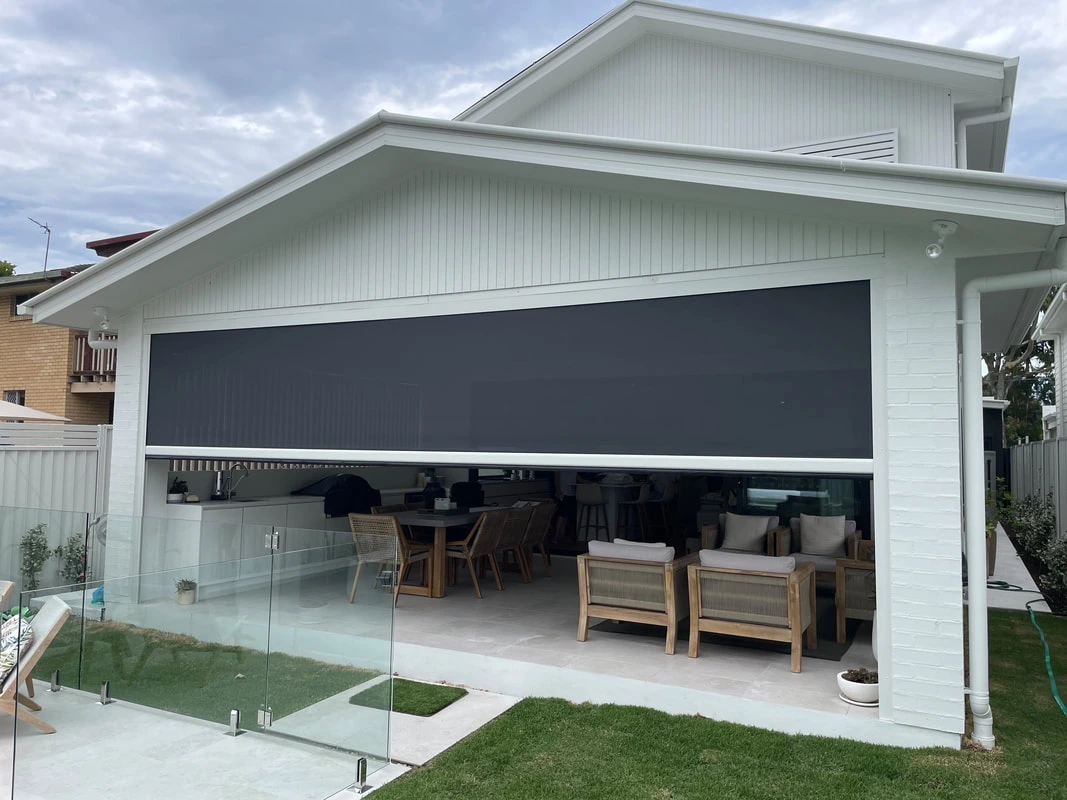 A wide charcoal zip-track outdoor blind partially raised over a large open alfresco entertaining area with timber and rattan furniture, glass pool fencing in the foreground.
