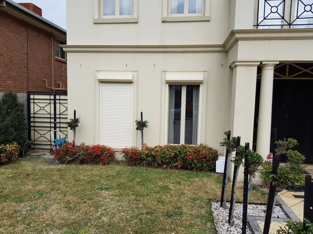 Two white roller shutters installed on the ground floor windows of a two-storey cream rendered home with classical architectural detailing, garden beds and lawn in the foreground.