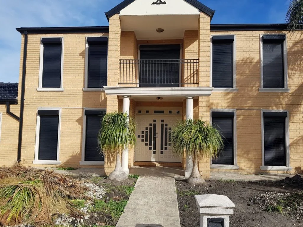 A large yellow brick two-storey home with charcoal roller shutters fully closed across all windows on the front facade, flanked by ponytail palm trees at the entrance.