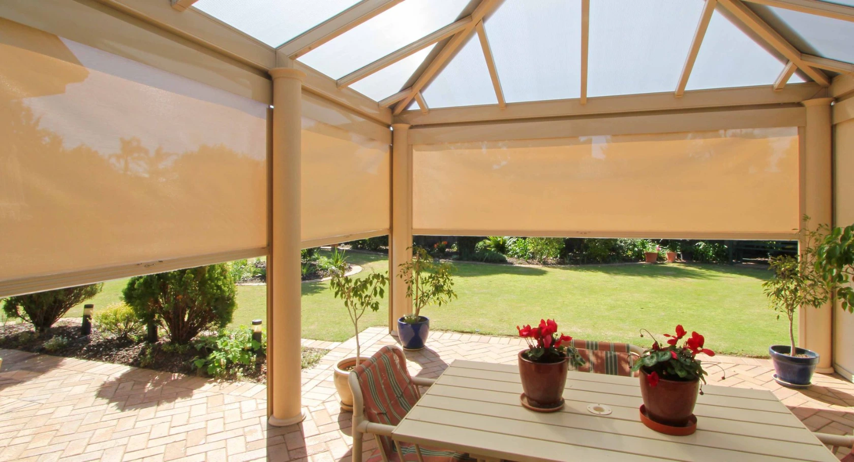 Cream slide-track outdoor blinds partially lowered around a gabled glass-roof patio enclosure, with a garden dining setting, potted flowers, and a lush lawn beyond, photographed from inside.