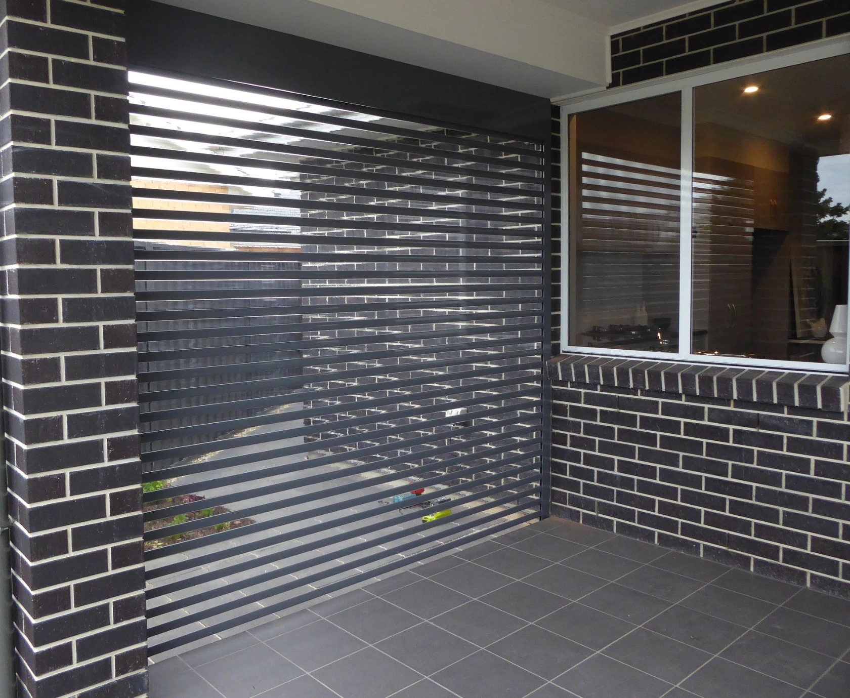 A charcoal clearview louvred roller shutter fully lowered at the entry of a dark brick home porch, with a sliding window visible beside it and tiled floor below.