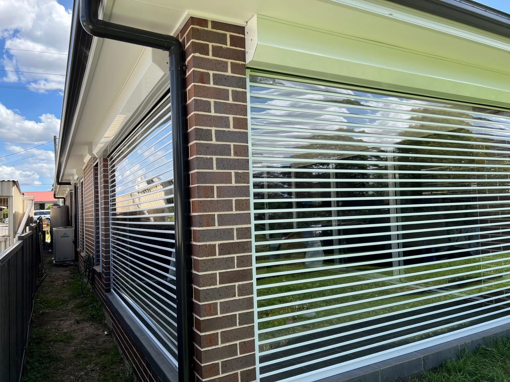 White clearview louvred roller shutters spanning the full side façade of a red brick home, reflecting sky and garden in the glazed slats, with a downpipe and air conditioner visible.