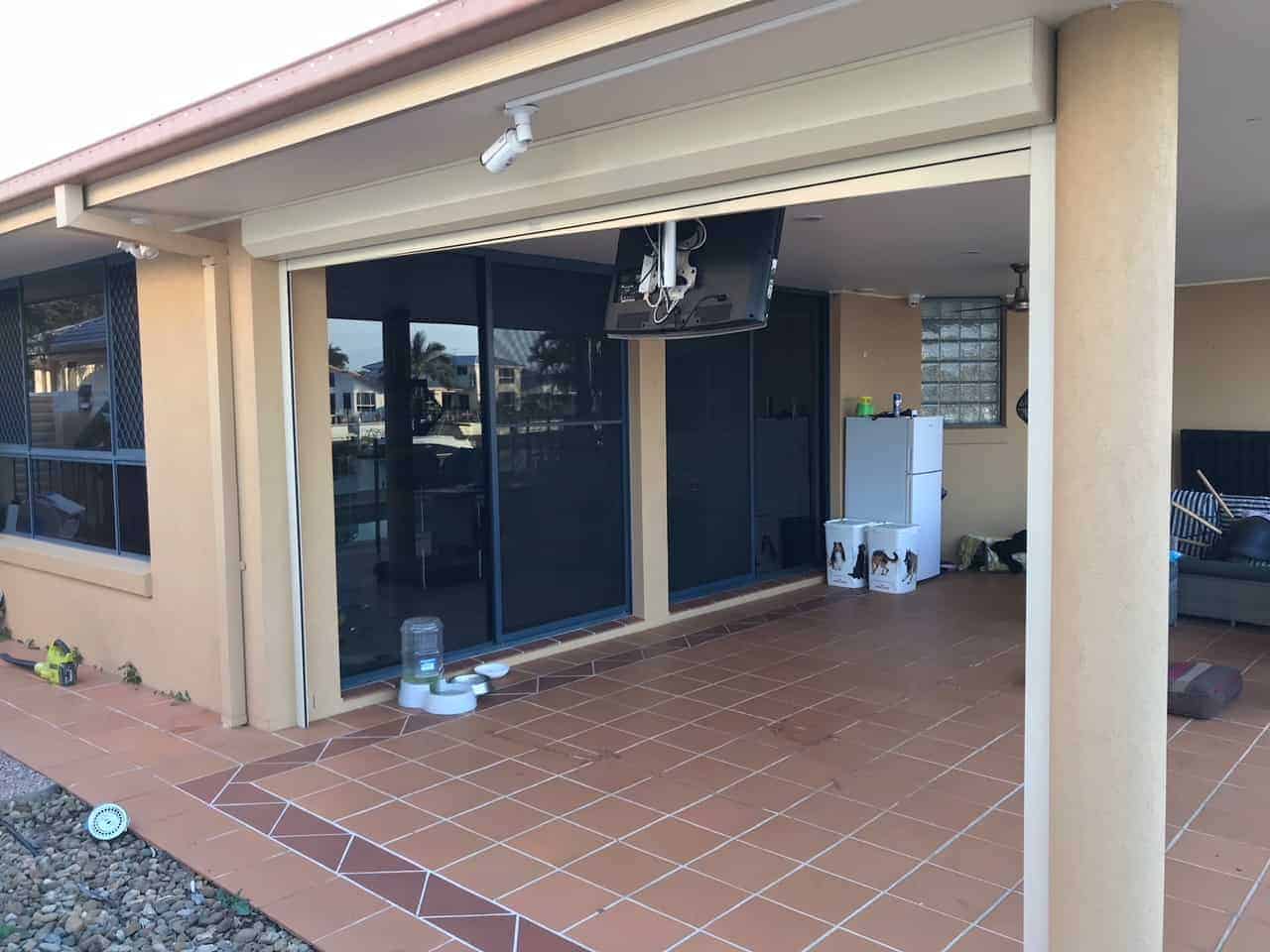 A cream roller shutter fully closed over a wide angled bay window on a red brick home, set in a burgundy aluminium frame with an agave plant and gnome ornament in the foreground.