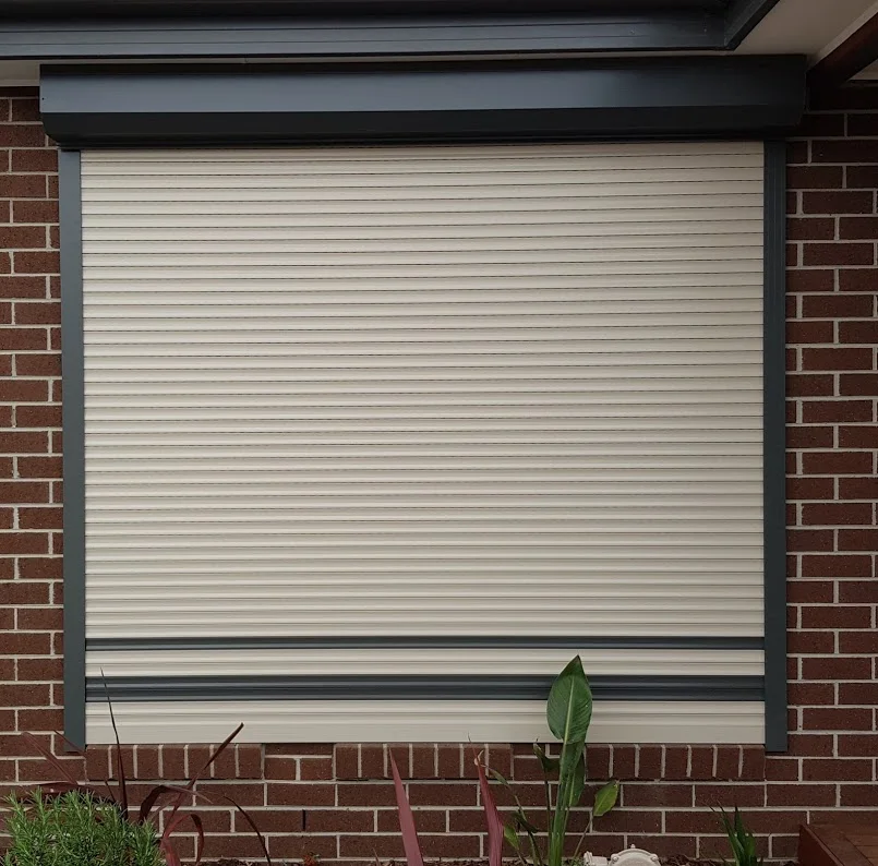 A single beige roller shutter installed over a large corner window on a brick veneer home, with a satellite dish visible on the roof.