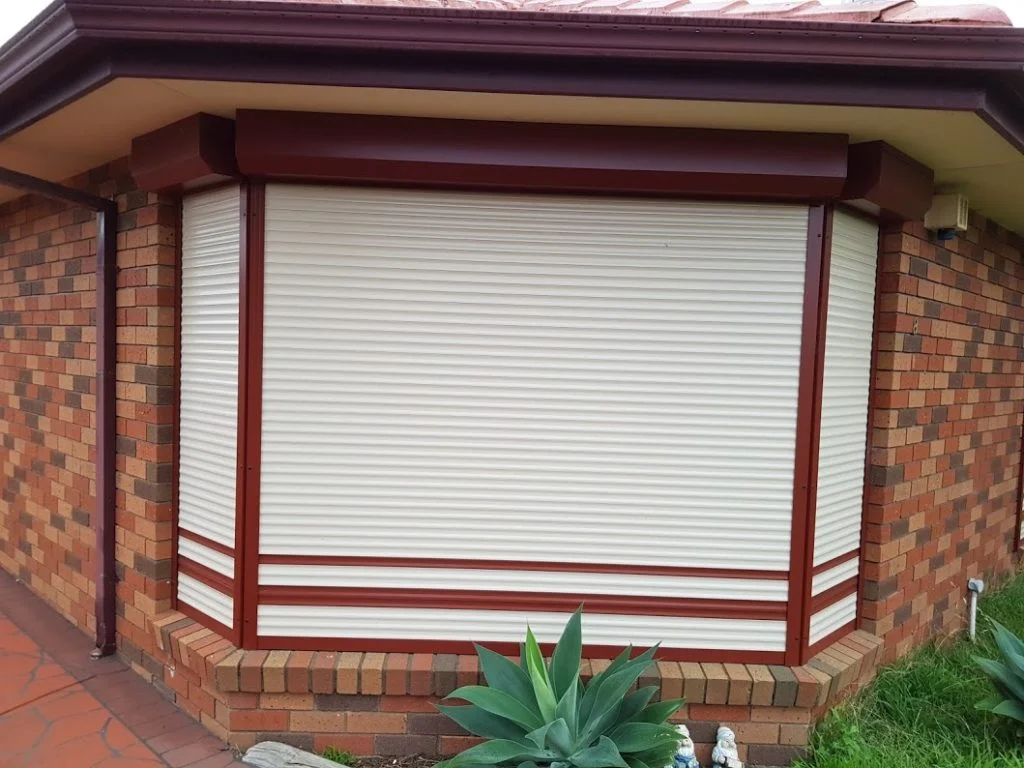Two charcoal roller shutters installed on the ground floor of a modern two-storey dark brick home, with an exposed aggregate driveway and glass balustrade balcony above.