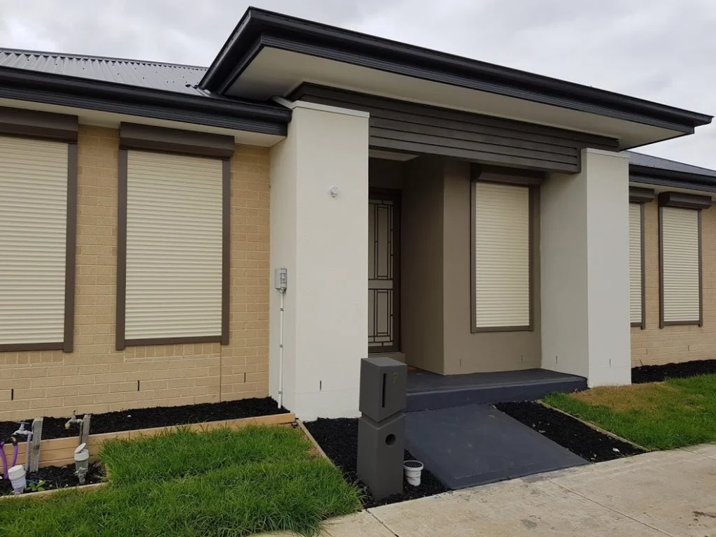 Multiple cream roller shutters fully closed across all windows on the front facade of a single-storey cream brick home with a dark roof, photographed from the street.