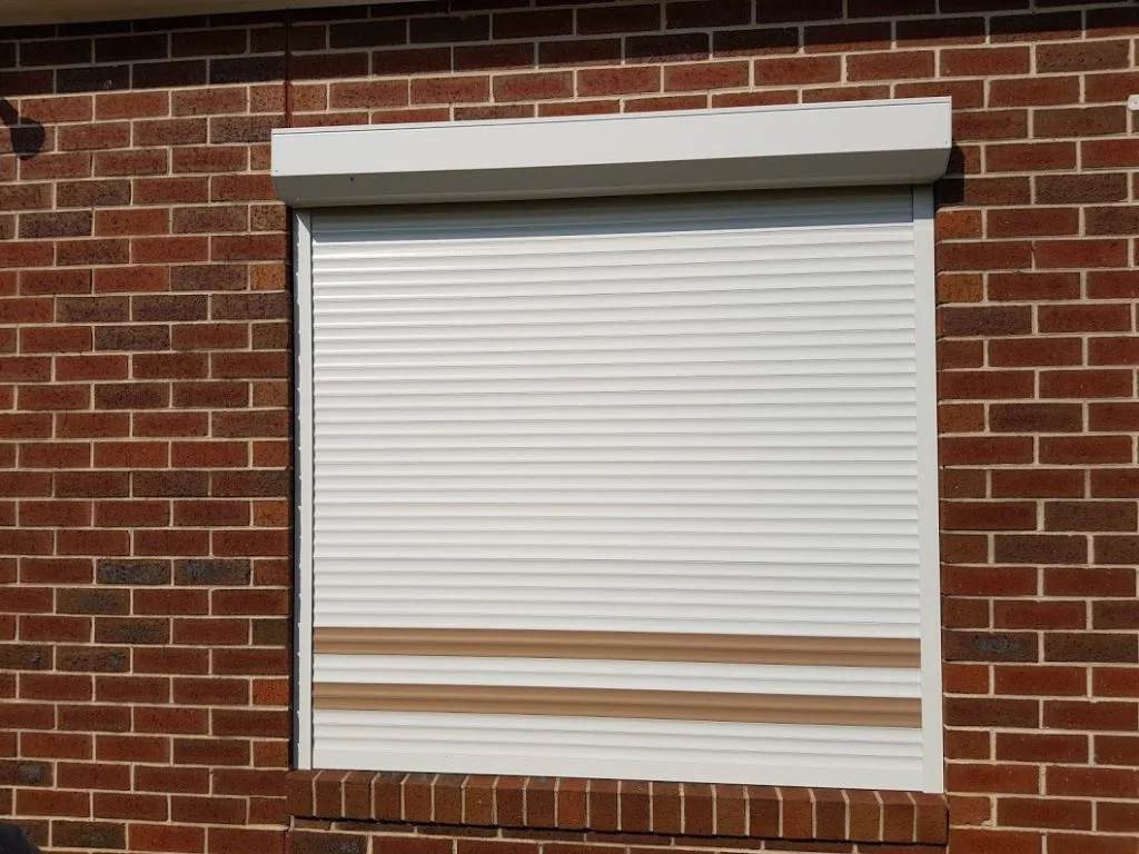 Two charcoal roller shutters installed on the ground floor of a modern two-storey dark brick home, with an exposed aggregate driveway and glass balustrade balcony above.