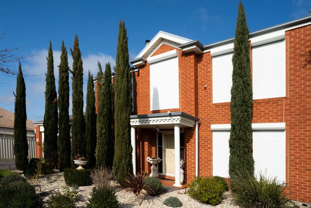 Three white roller shutters fully closed across the upper and lower windows of a two-storey red brick home with classical detailing, flanked by tall Italian cypress trees against a blue sky.