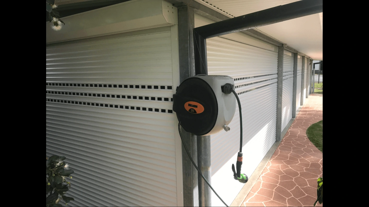 Multiple white perforated roller shutters fully closed along a covered alfresco walkway, with a retractable garden hose reel mounted on a steel post and terracotta paving visible to the right.