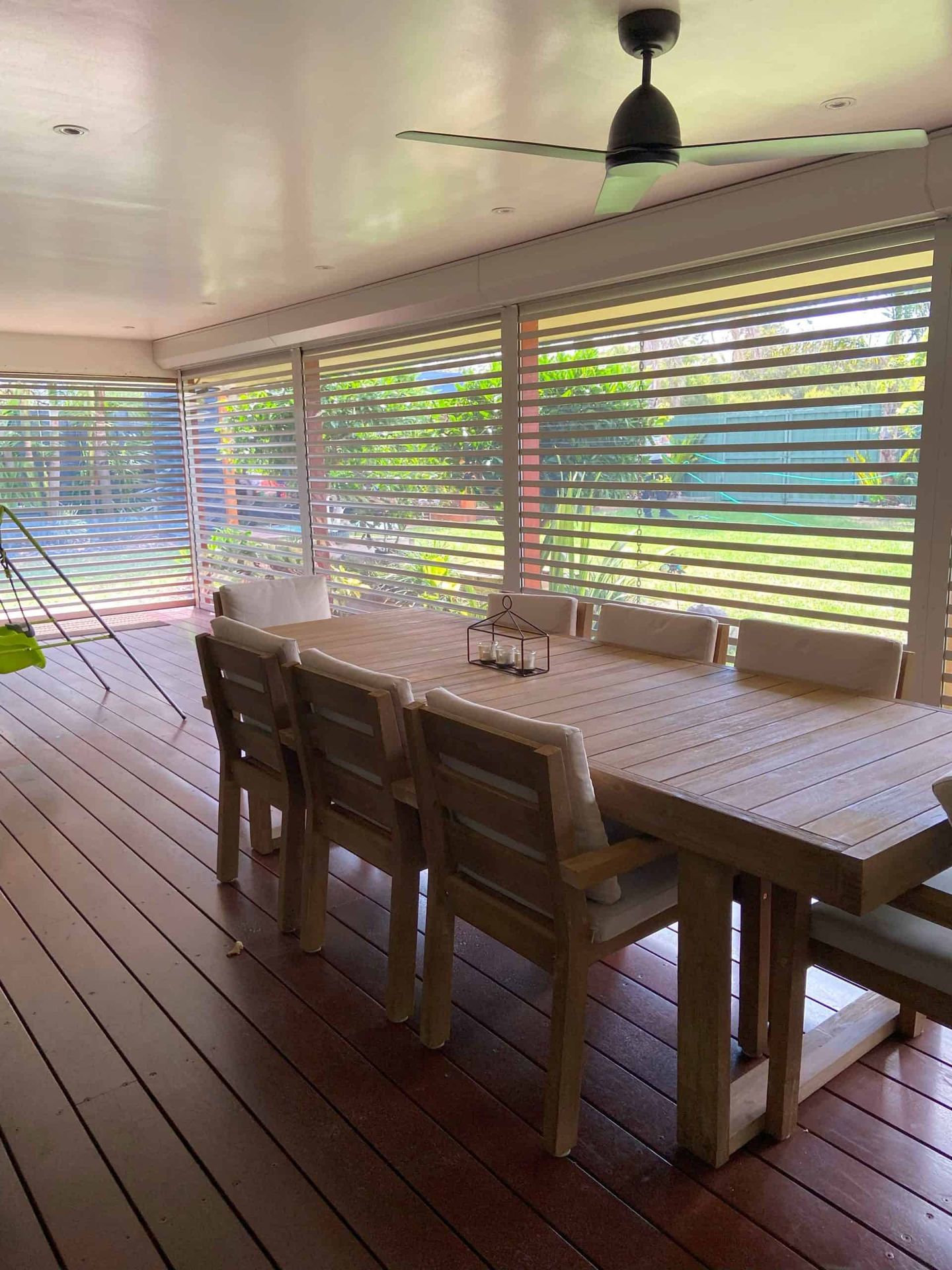 Clearview louvred roller shutters spanning the full width of a covered timber deck alfresco, allowing garden light to filter through, with a ceiling fan above and timber dining table below.