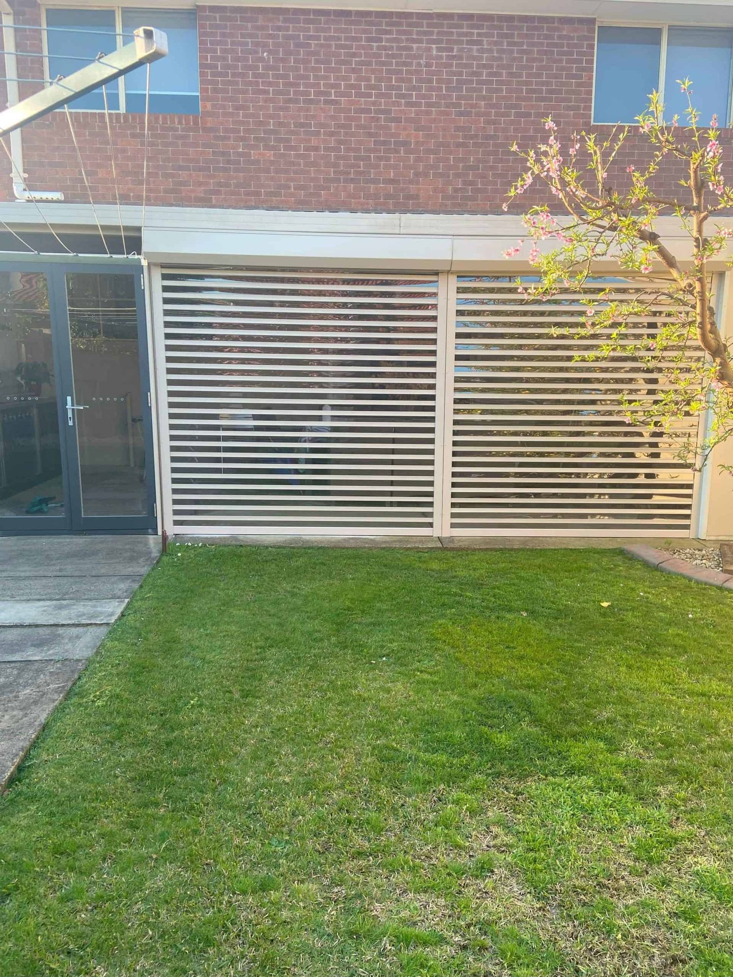 Two white clearview louvred roller shutters fully lowered across a rear patio of a red brick home, with a flowering tree and lawn in the foreground.
