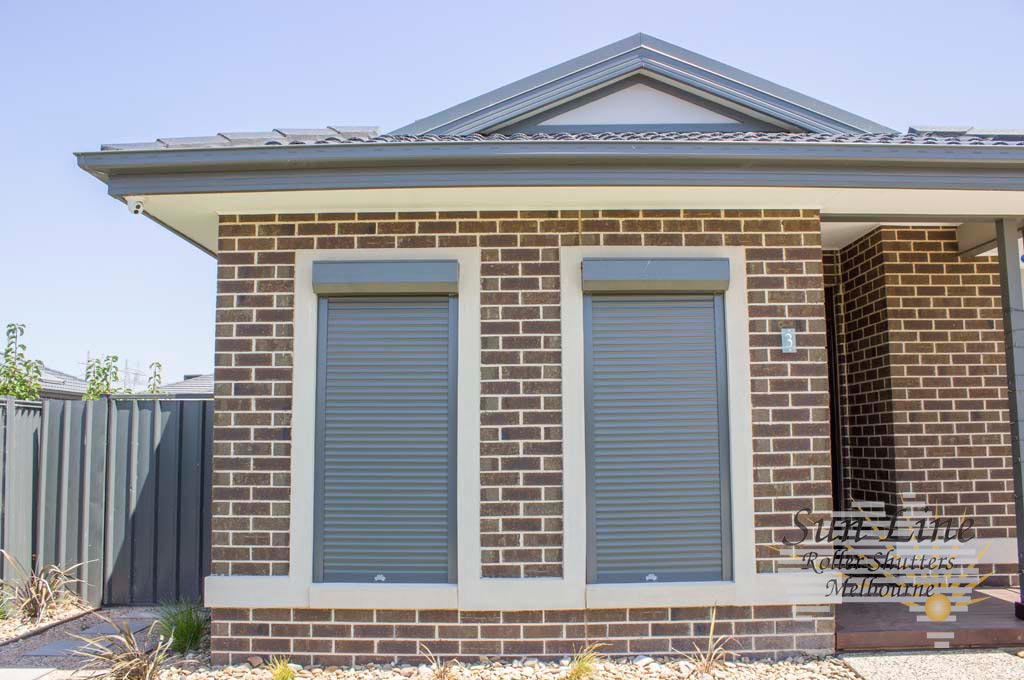 Three grey roller shutters fully closed over windows on the front of a red brick home, with ornamental grasses and a gravel garden in the foreground.