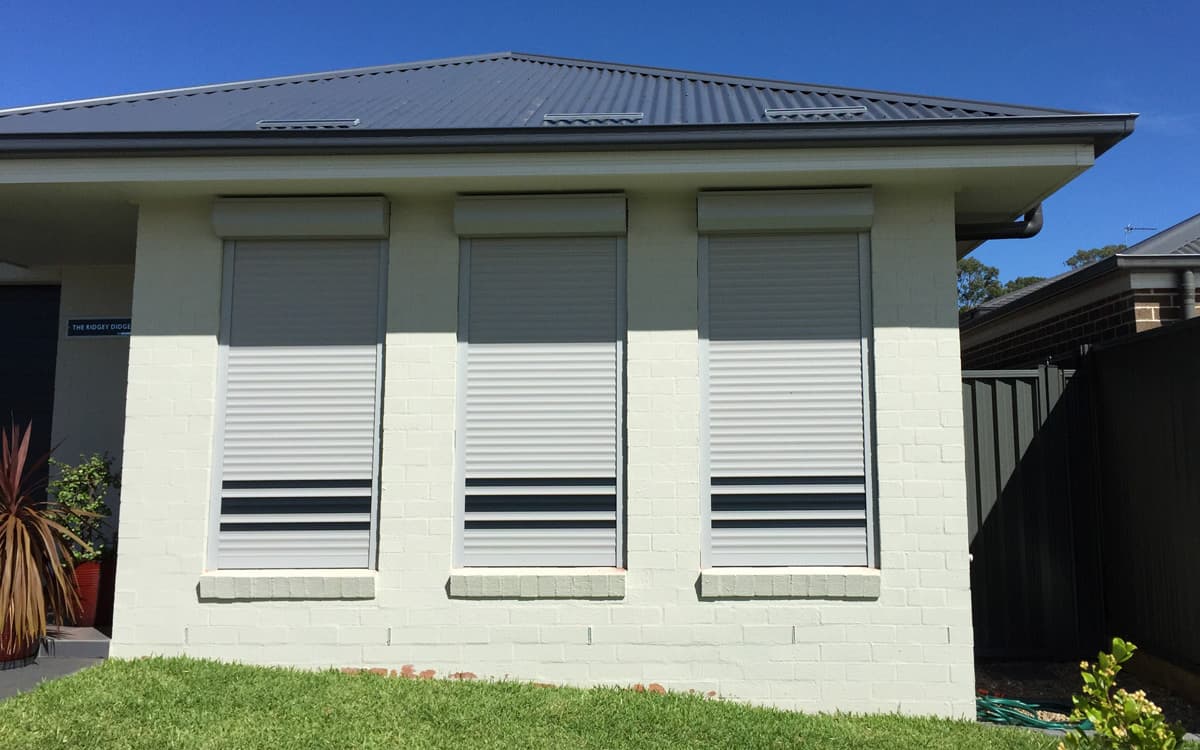 Three white roller shutters partially closed over windows on a rendered brick home exterior, with dark grey accents and a metal roof, set against a clear blue sky.