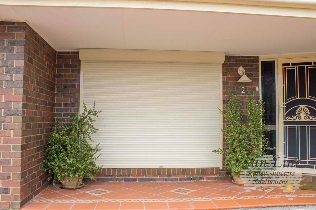 A cream-coloured roller shutter fully closed over a wide front window of a red brick home, flanked by potted shrubs on a tiled porch.