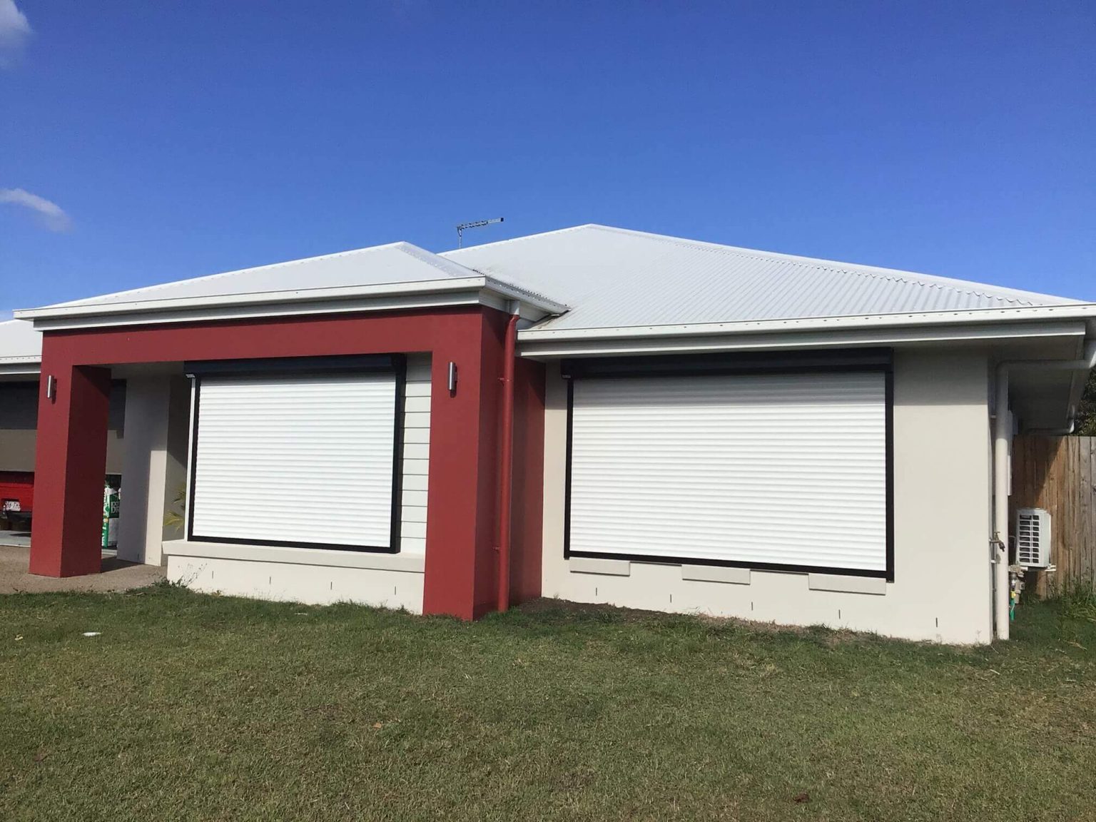 Two white roller shutters fully closed over large windows on a rendered cream and red feature-wall home, with dark charcoal frames and a lawn in the foreground under a blue sky.